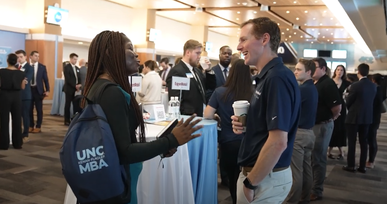 A female student with backpack speaks to an older male from a company during an MBA recruiting event.